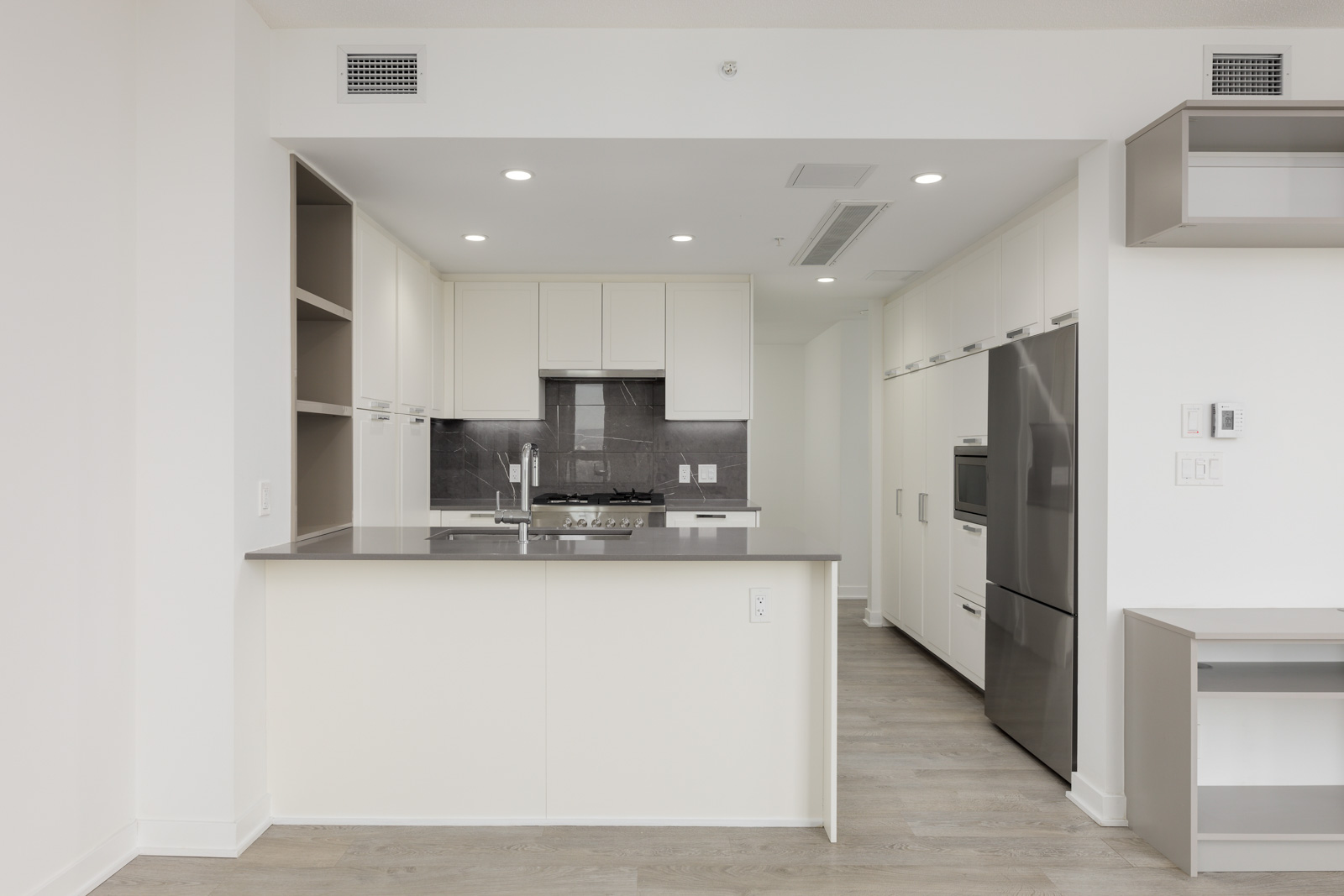 Modern kitchen with white cabinets, gray countertops, and stainless steel appliances, including an oven and fridge. Recessed lighting and gray backsplash complete the design.