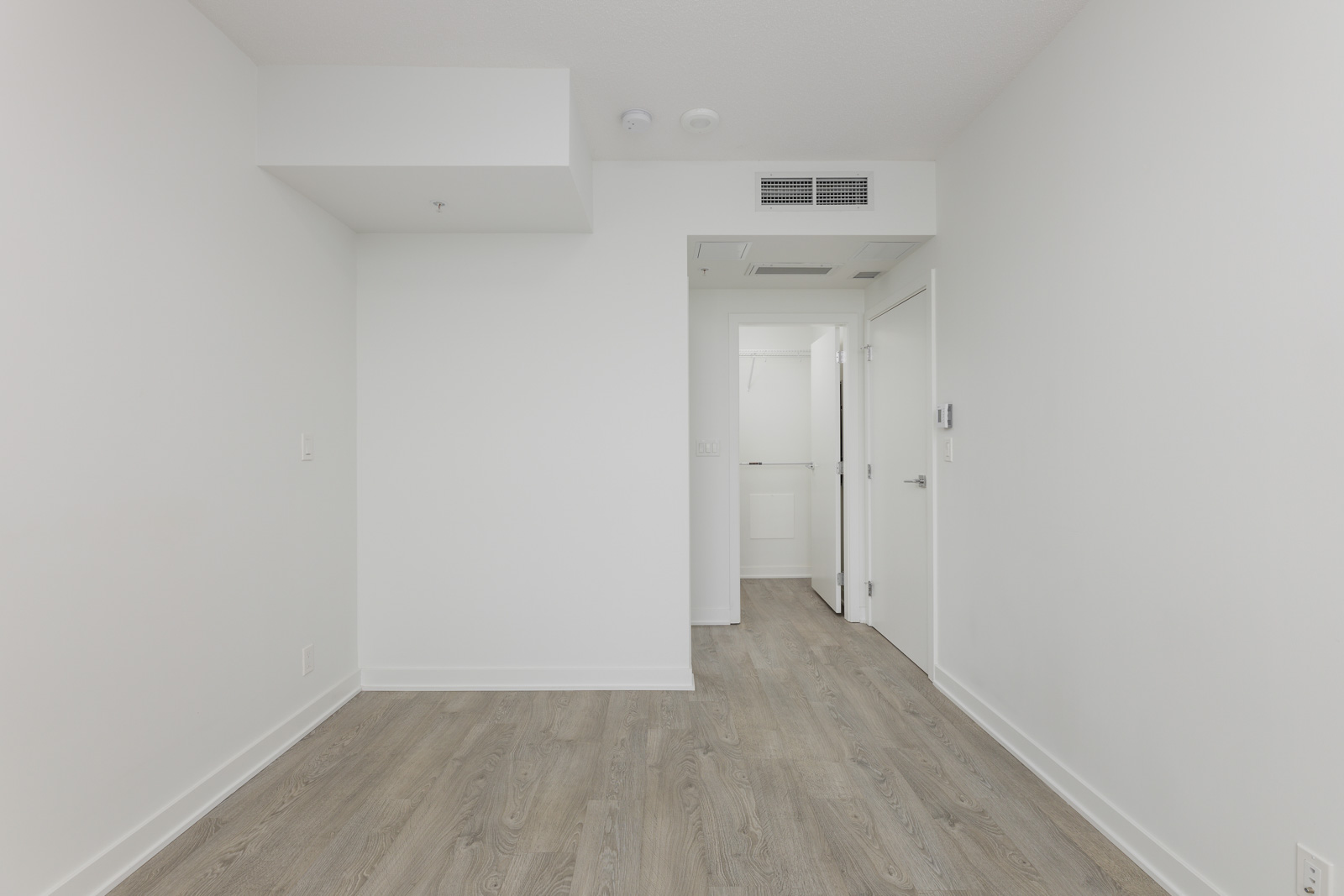 Empty white room with wood flooring, a vent on the ceiling, and a doorway leading to a hallway. Walls and ceiling are plain with no decorations.