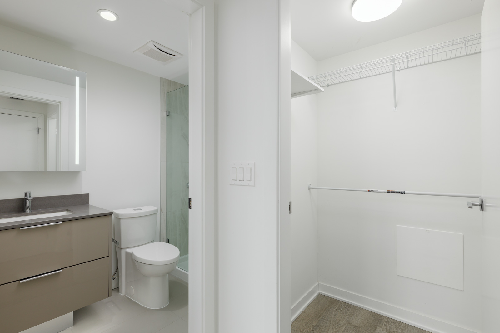 Modern bathroom with a sink, vanity, toilet, and shower, connected to a walk-in closet with a wooden floor and wire shelf.