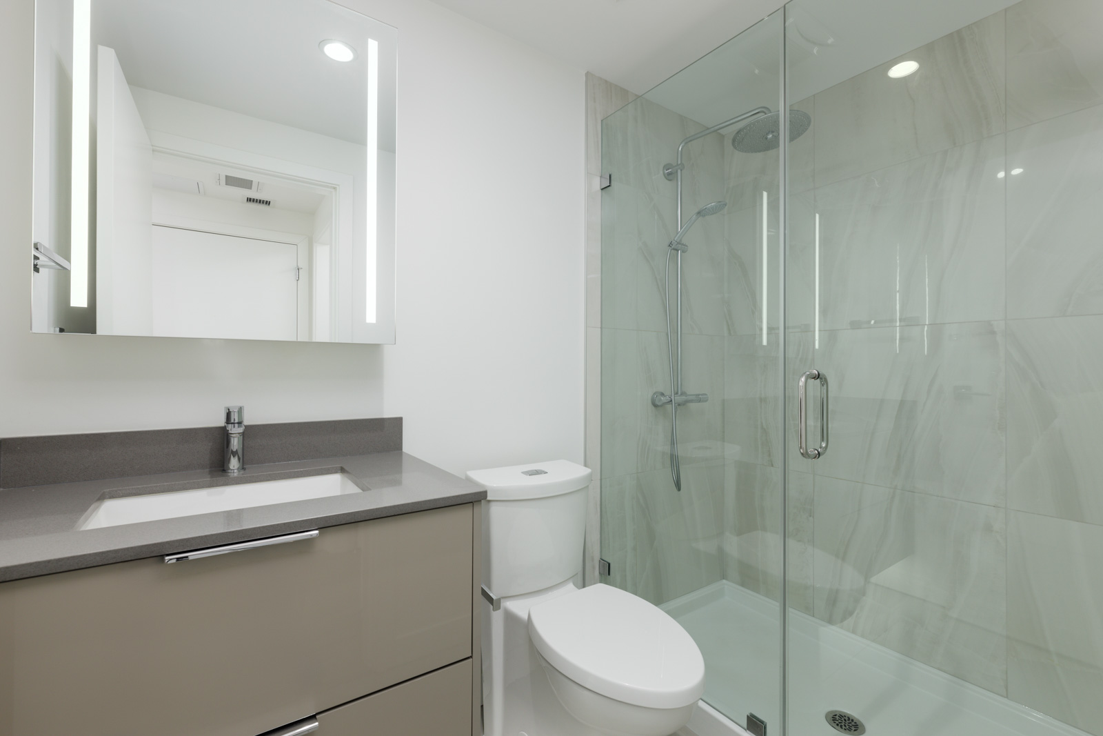 Modern bathroom with a glass-enclosed shower, gray countertop sink, and toilet. Walls are white, and there is a mirrored cabinet above the sink.
