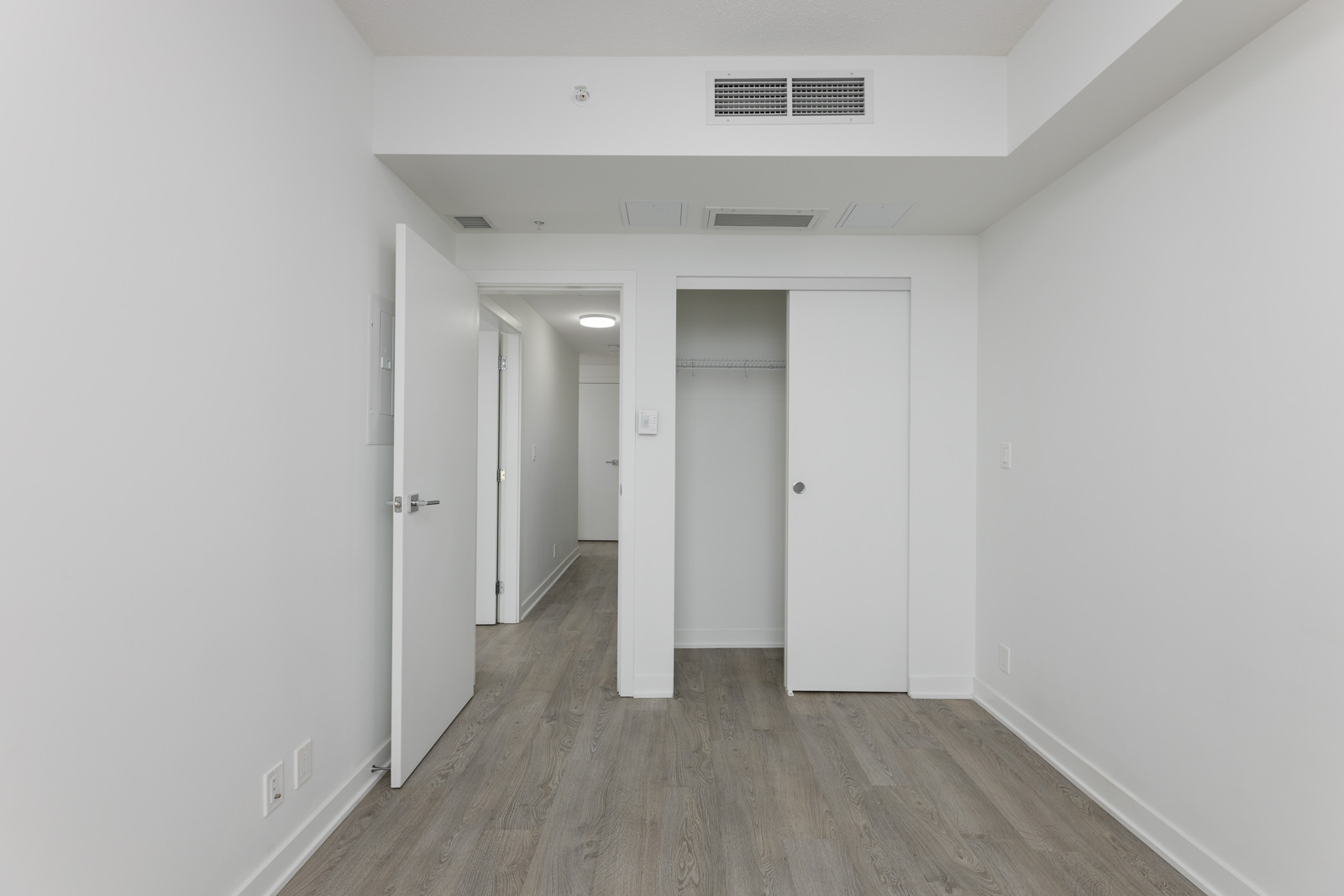 Empty room with white walls, wooden floor, and an open door. Closet with sliding doors on the right. Ceiling lights and a visible hallway.