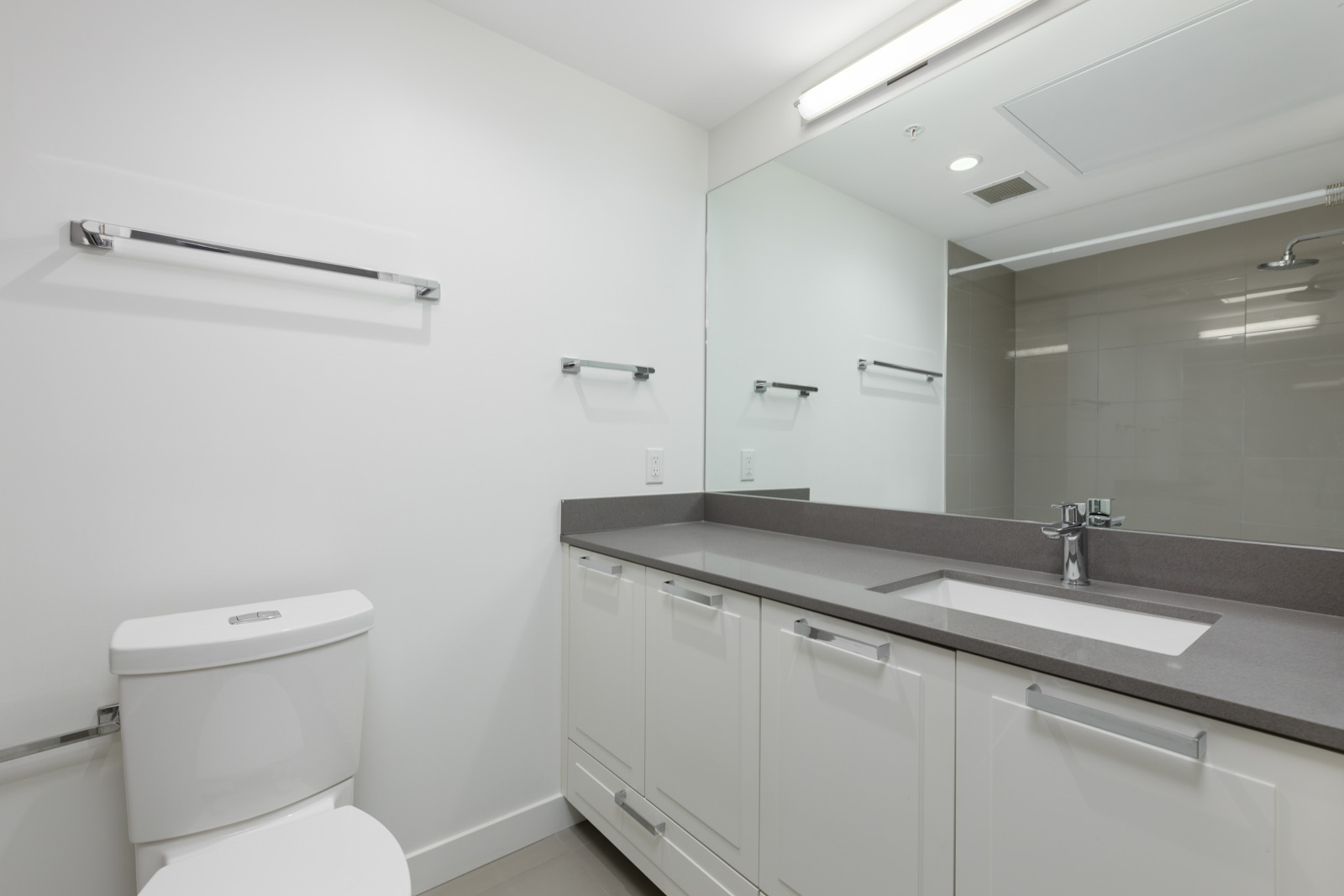 Modern bathroom with white cabinetry, a large mirror, sink, and a toilet. There are towel racks on the wall and a shower area with a glass door.