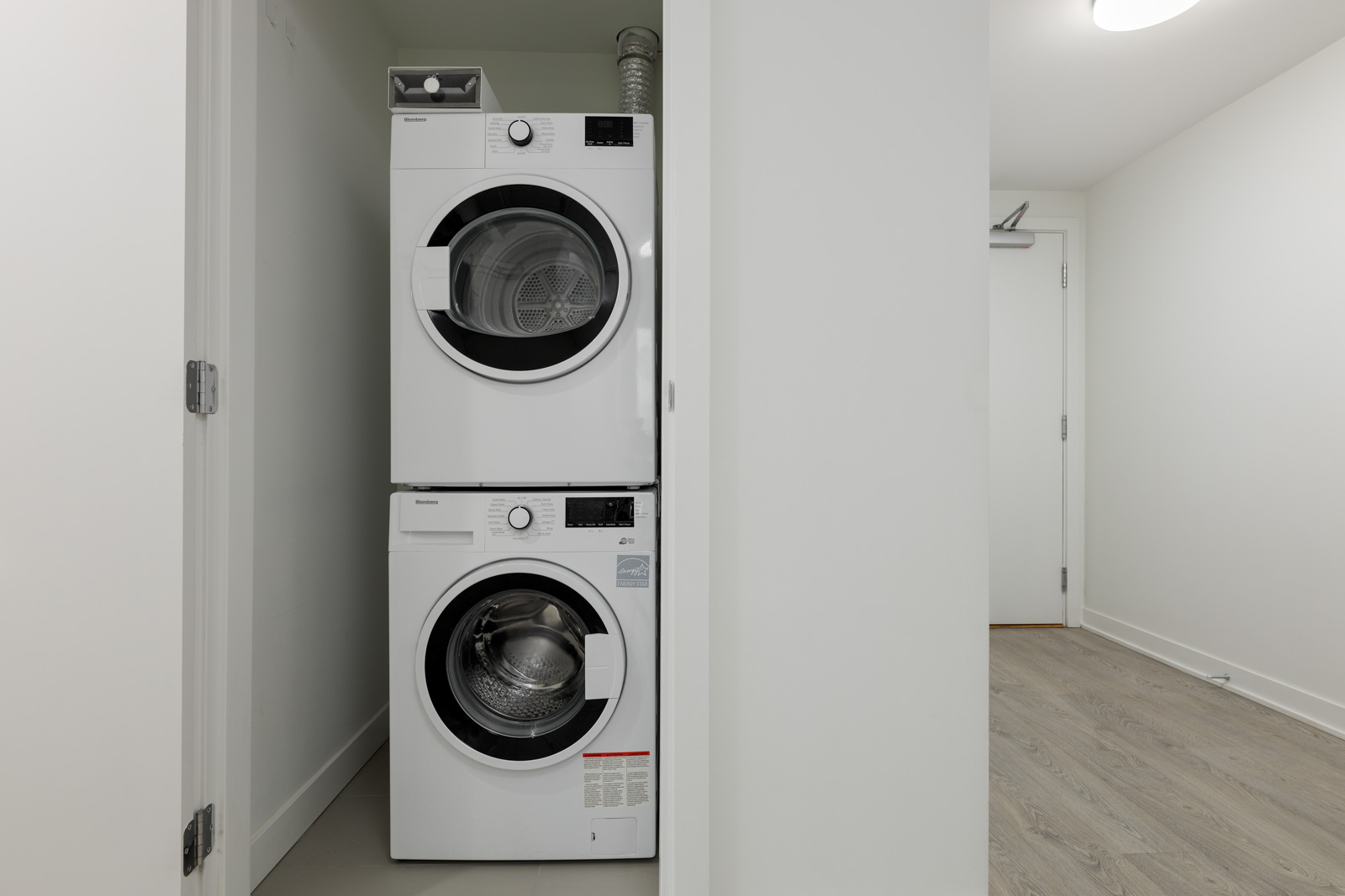 Stacked washer and dryer in a white closet, with a wooden floor and a closed door in the background.