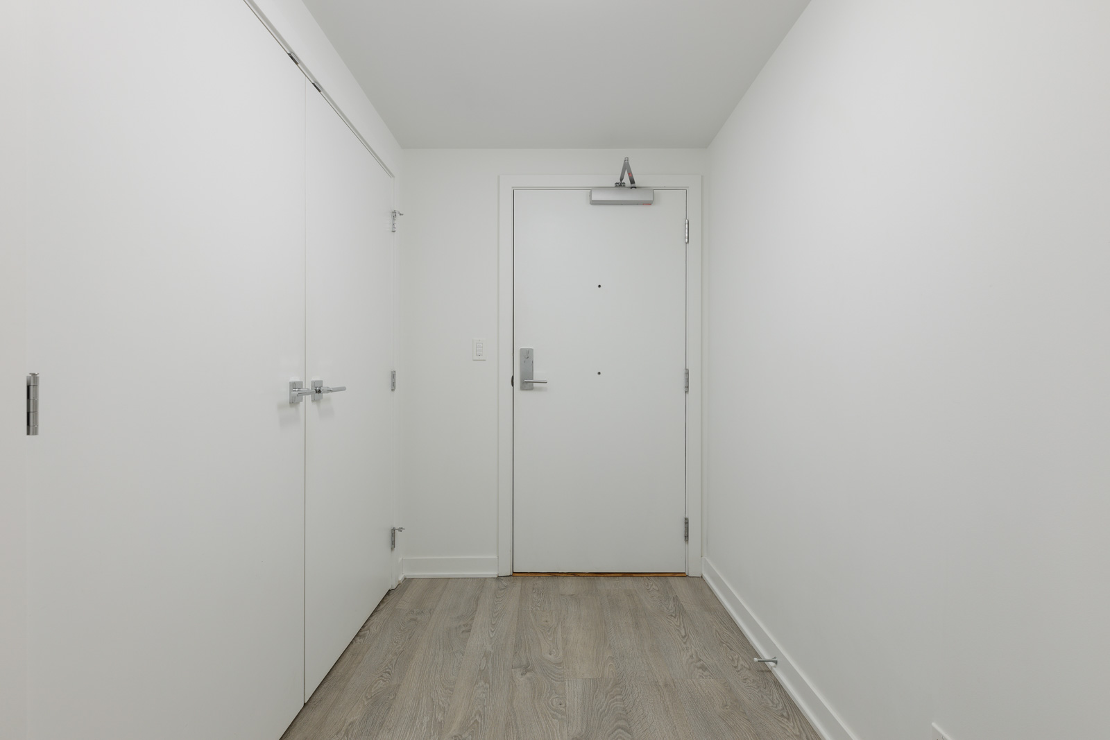 Minimalist interior with a white door, two closet doors on the left, and light wooden flooring. Walls and ceiling are white.