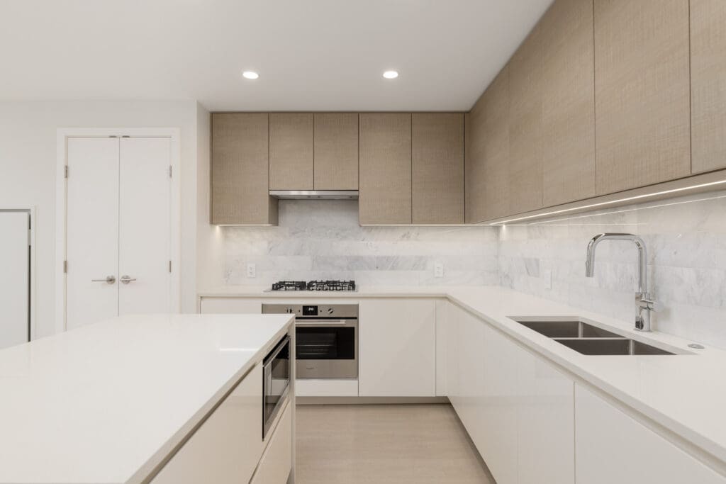 Modern kitchen with white cabinets, marble backsplash, and wooden upper cabinets. Includes a built-in oven, sink, and kitchen island.