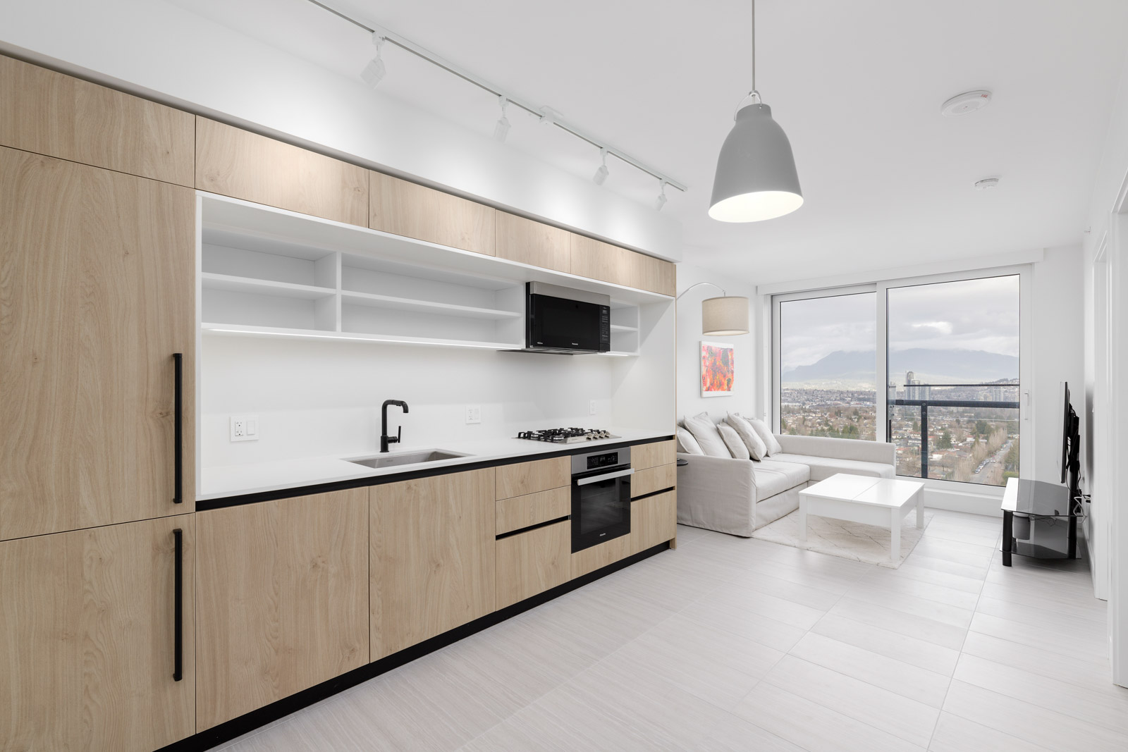 Minimalist kitchen and living room with light wood cabinets, white walls, sofa, and a large window with a city view.