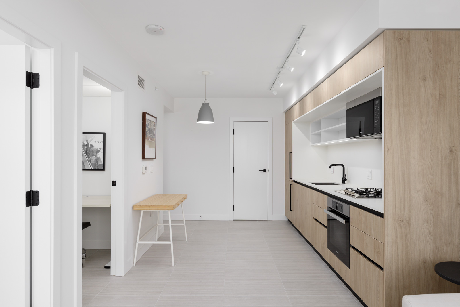 Minimalist kitchen with light wood cabinets, black appliances, and a white countertop. Features a small table with a bar stool and wall-mounted lighting. White walls and tiled floor.