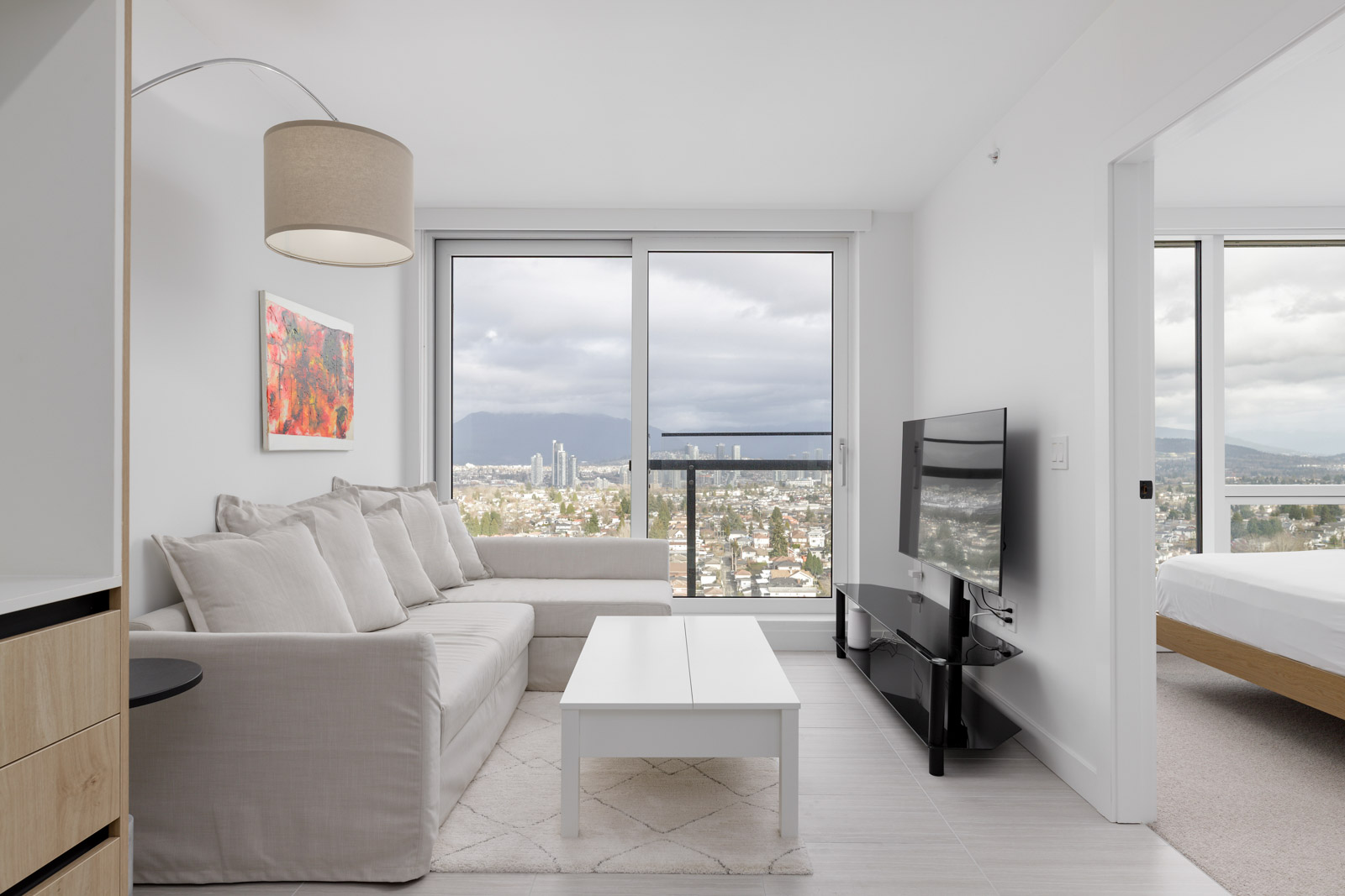Modern living room with a gray sofa, wall-mounted TV, and large windows overlooking a cityscape. Bedroom with a bed is visible through an open sliding door.