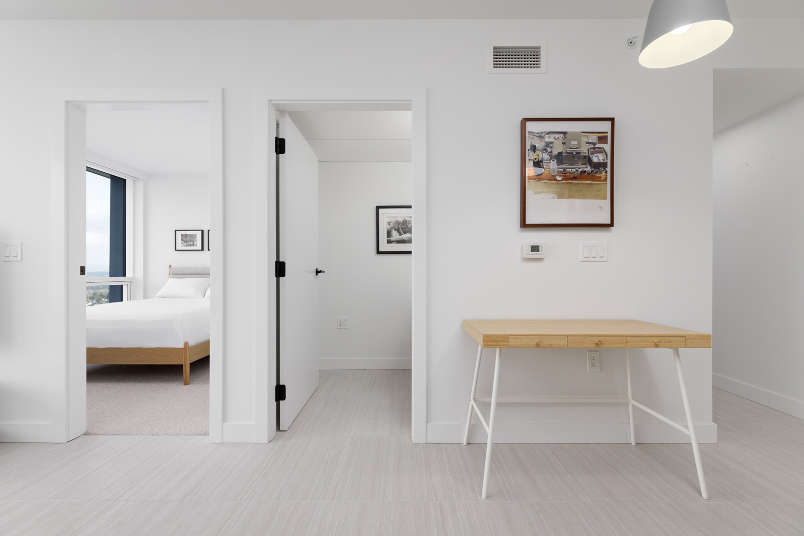 Minimalist room with a wooden desk, a doorway leading to a bedroom with a bed, and framed photos on white walls.