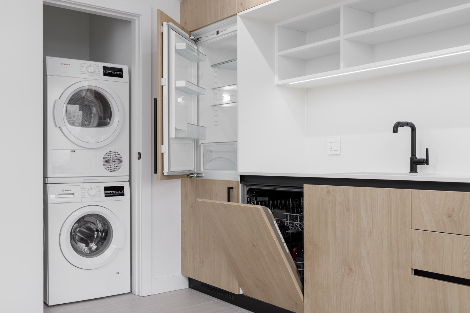 Modern laundry and kitchen area with a stacked washer and dryer, open refrigerator, dishwasher, and wooden cabinets under a black faucet.