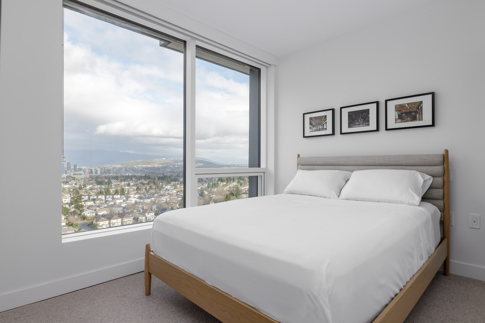 A minimalist bedroom with a bed, large window, and framed pictures on a white wall. A cityscape view is visible through the window.
