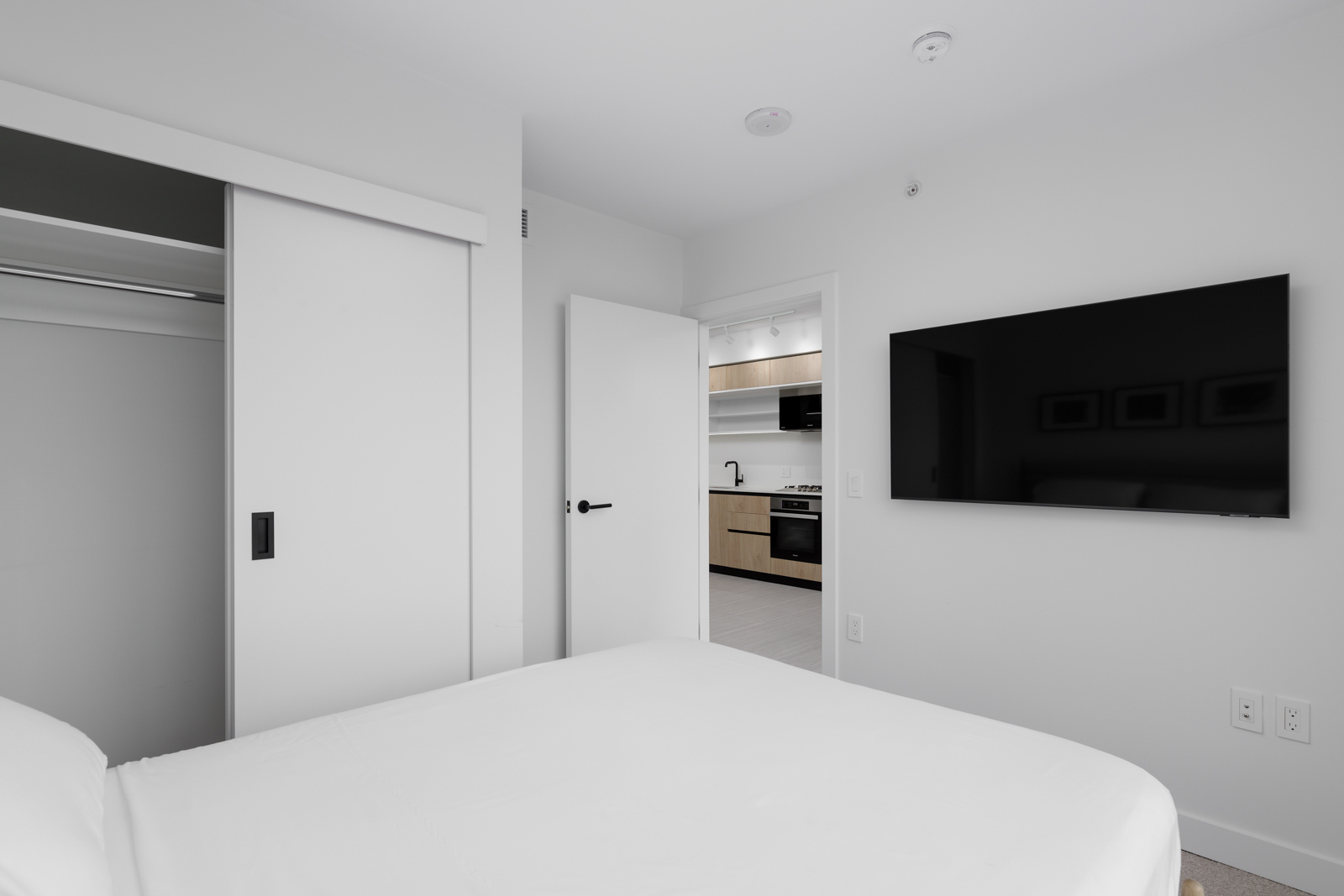 A minimalist bedroom with a white bed, wall-mounted TV, open closet, and view into a kitchen with light wood cabinetry.