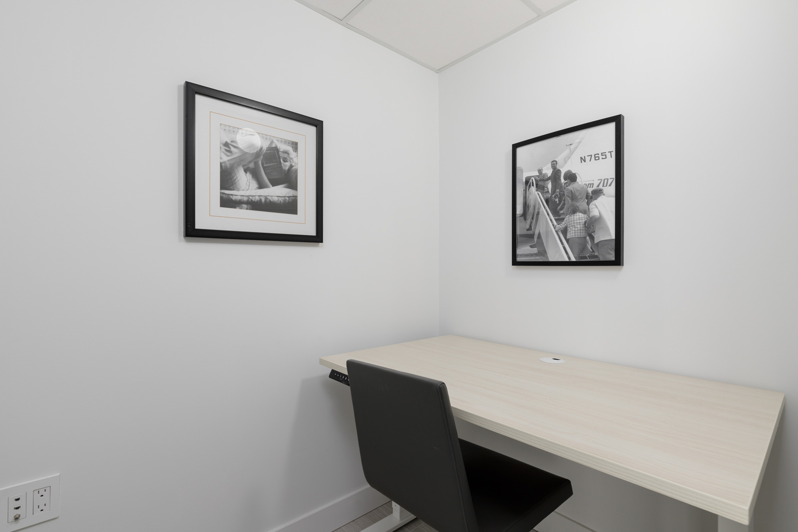 Minimalist office corner with a wooden desk and black chair, featuring two black-and-white framed photographs on white walls.