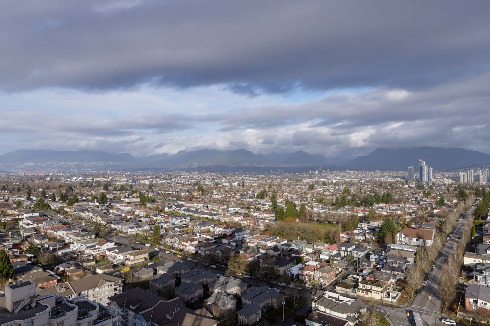 Aerial view of a cityscape with rows of residential houses, a few tall buildings, and distant mountains under a cloudy sky.