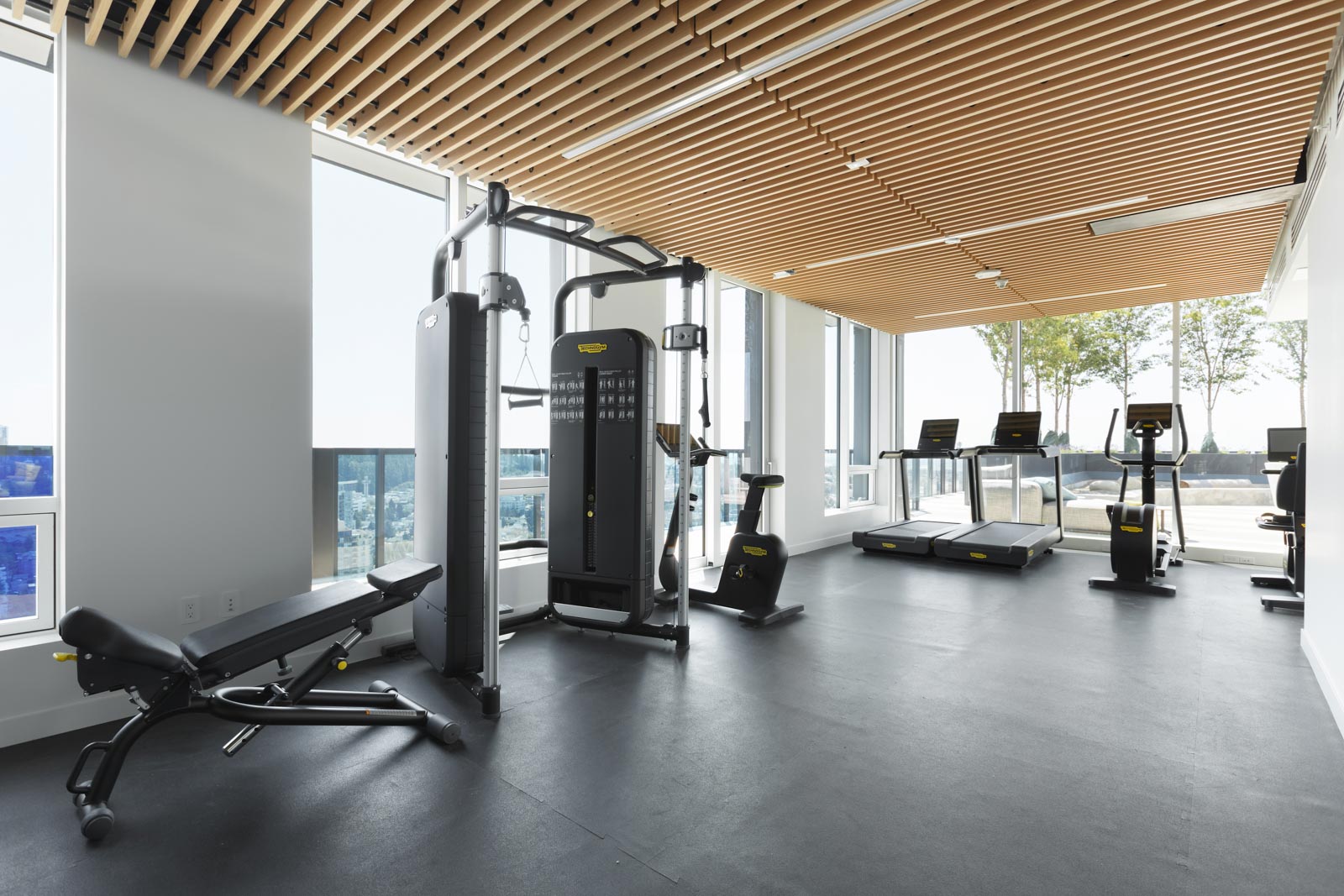 A modern gym with exercise equipment including a multi-station machine, stationary bicycles, treadmills, and a weight bench. Large windows offer a view outside.