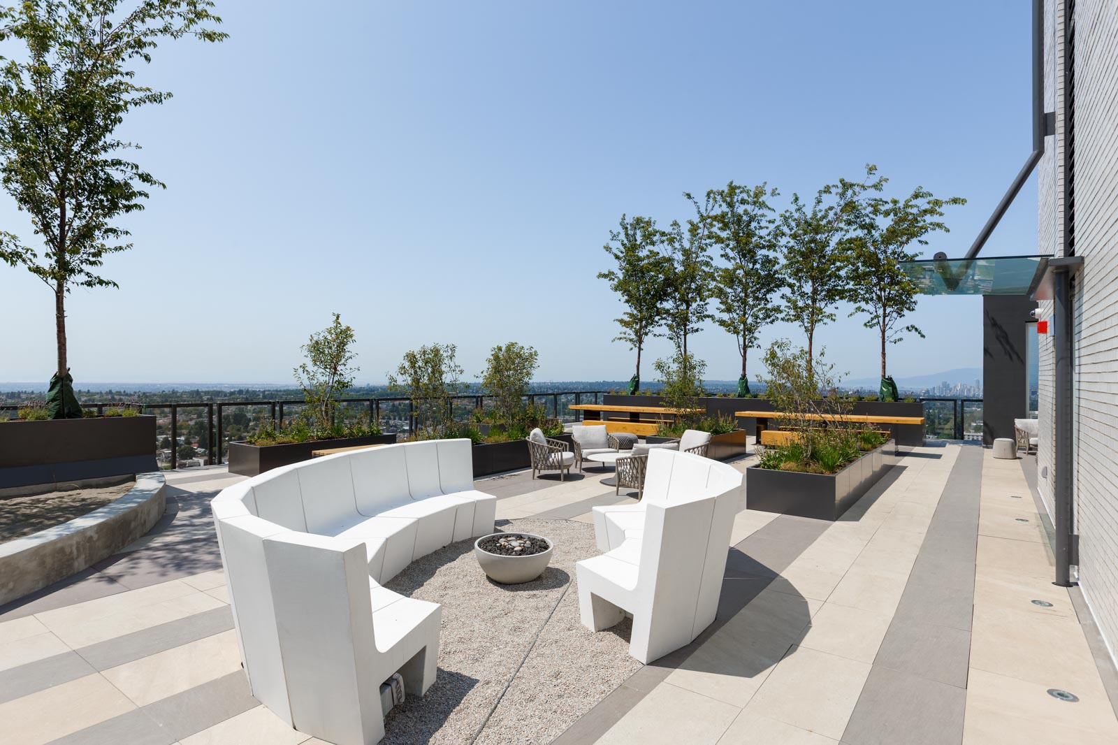 Rooftop terrace with white curved seating, fire pit, and planters. Trees and a city skyline are visible in the background.