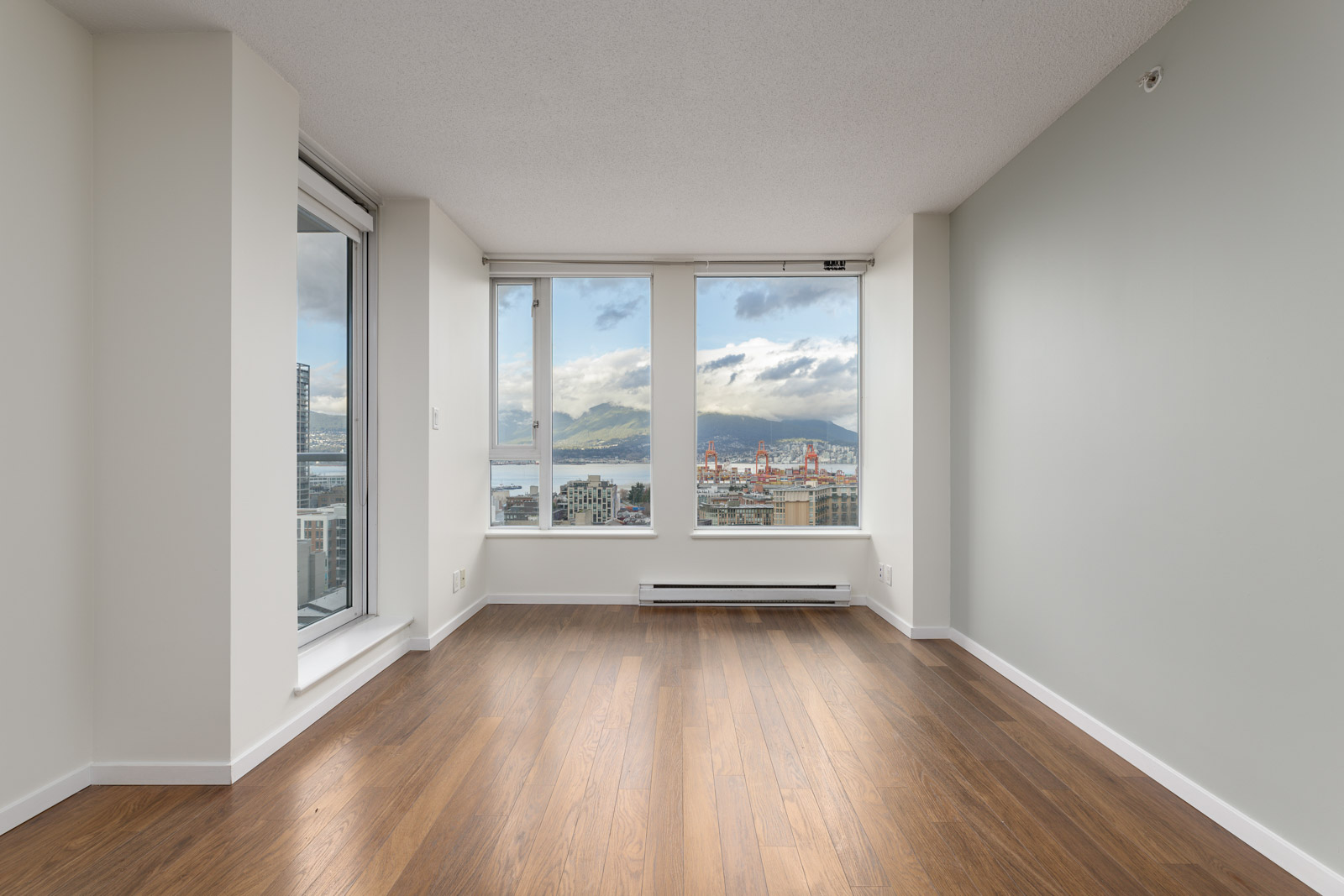 An empty room with wooden floors and large windows offering a view of city buildings and mountains in the distance.