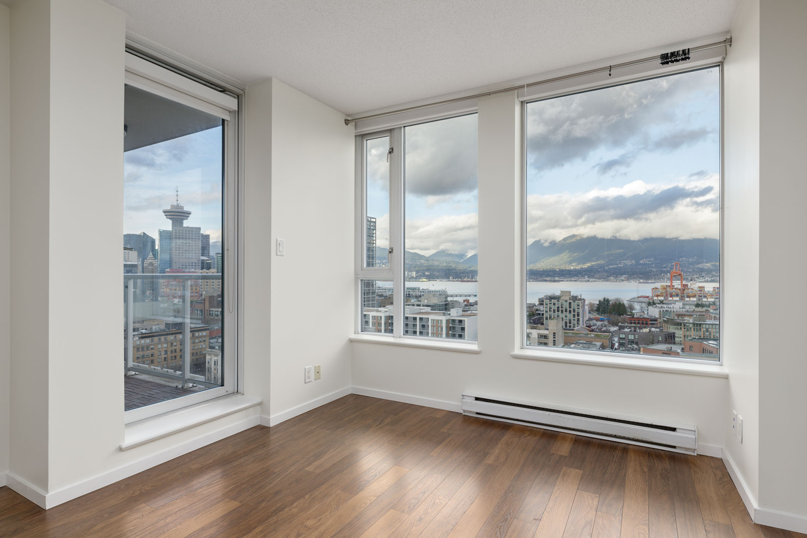 Modern apartment interior with large windows and a view of a cityscape, mountains, and sky. Hardwood floors and neutral walls are visible.