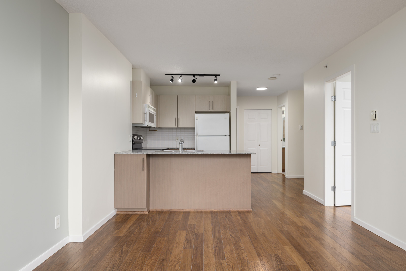 Modern kitchen area with light wood cabinetry, stainless steel appliances, and a track lighting fixture. The space features hardwood floors and white walls.