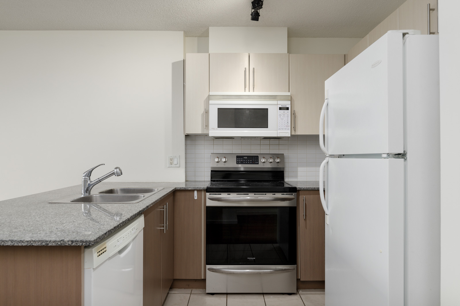 A modern kitchen with a granite countertop, stainless steel stove, white microwave, white refrigerator, and sink with a dishwasher. Cabinets are light wood and white.
