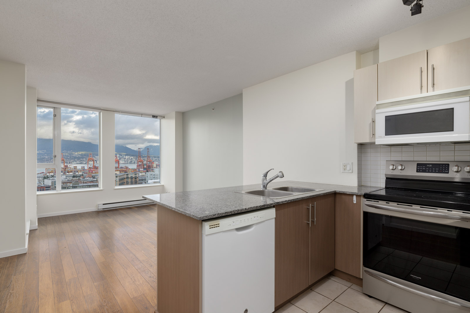 Modern apartment kitchen with granite countertops and stainless steel appliances, overlooking a spacious living area and large windows with a city and mountain view.
