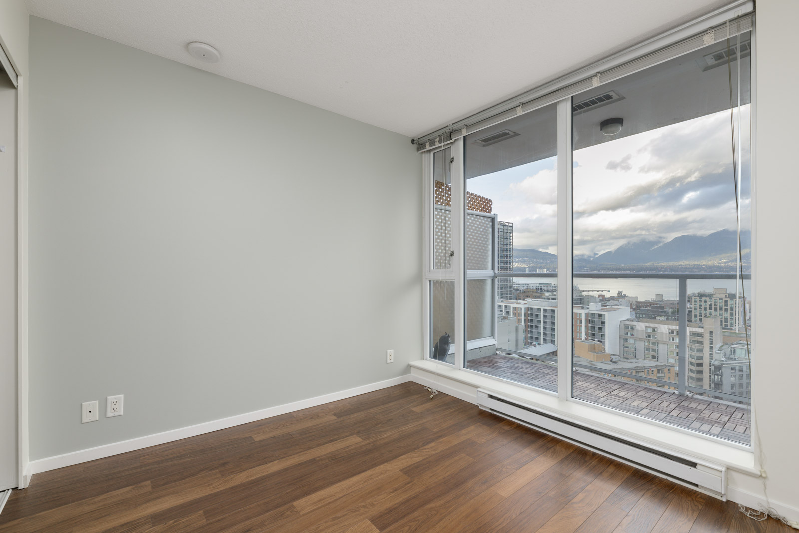 Empty room with light gray walls, hardwood floor, and large windows offering a view of a cityscape and mountains.