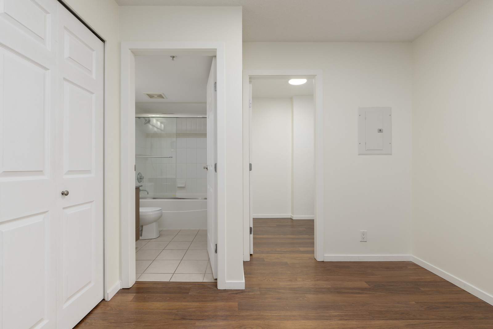 Empty room with wooden floor, white walls, and two open doors. One door leads to a bathroom with a tiled floor and bathtub; the other leads to a hallway with similar flooring.