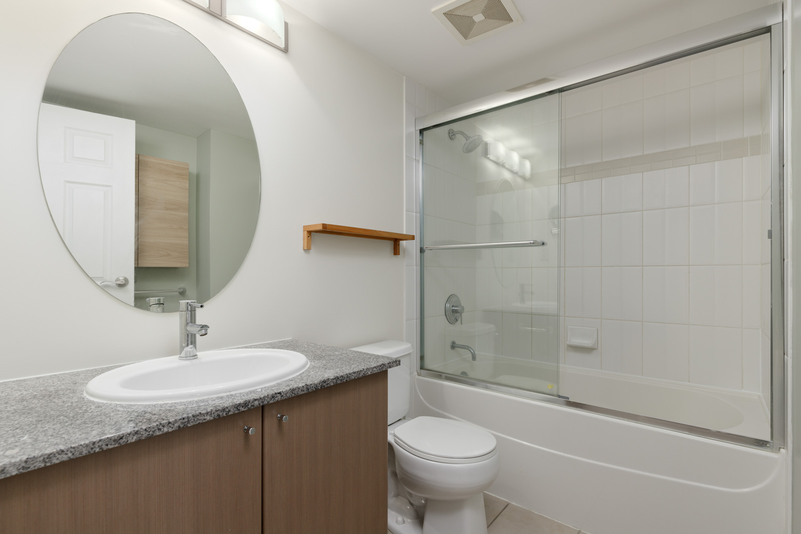Modern bathroom with oval mirror, granite countertop sink, wooden cabinet, and bathtub with glass shower doors. White tiled walls and a small wooden shelf above the toilet complete the look.