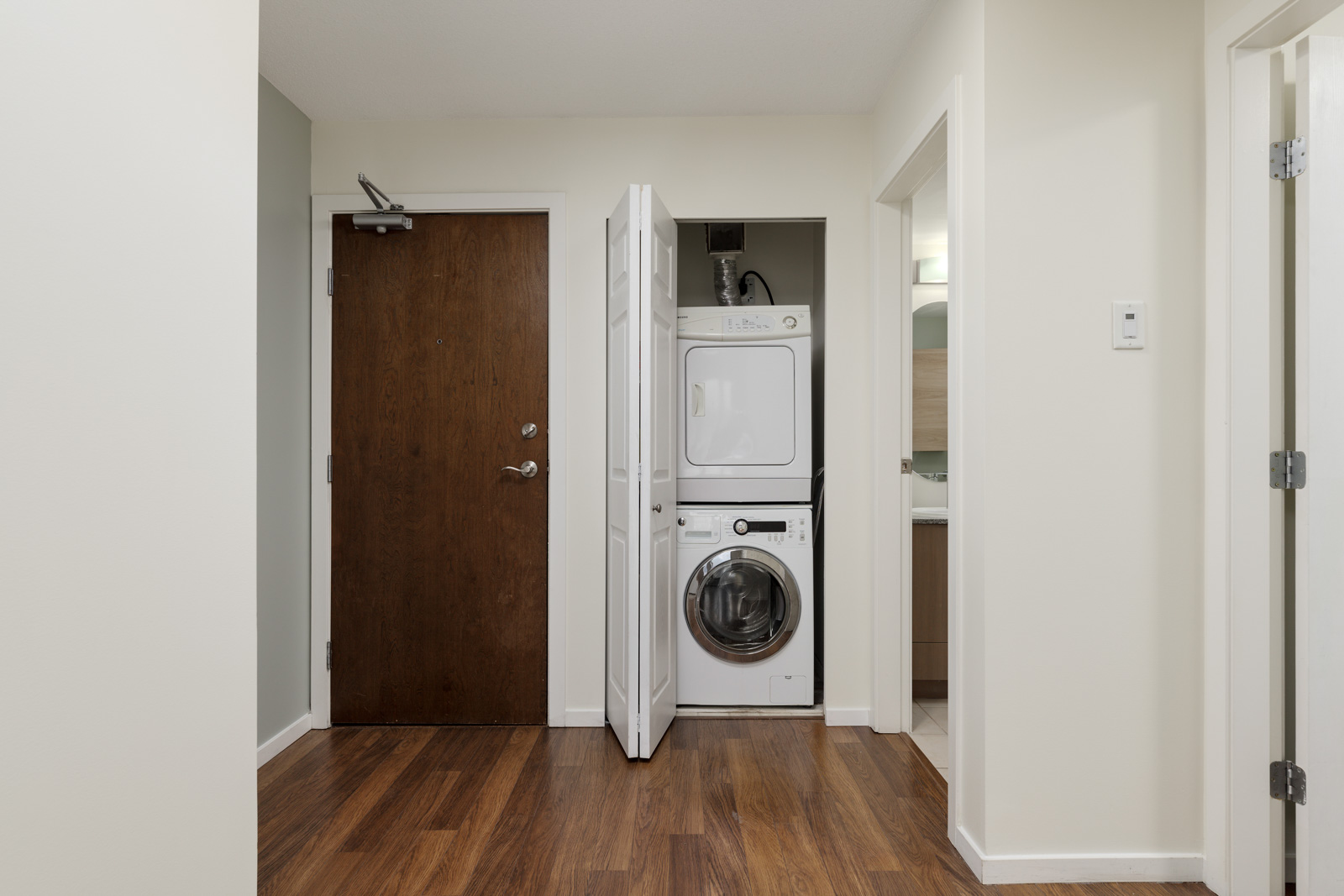 A compact laundry area with a stacked washer and dryer is situated in a closet with white doors, in a room with wood flooring and light-colored walls.