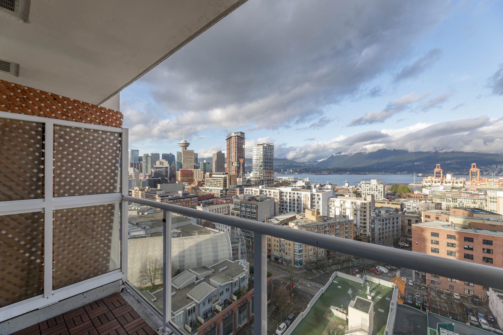 Cityscape view from a balcony overlooking downtown skyscrapers, a body of water, and distant mountains under a partly cloudy sky.