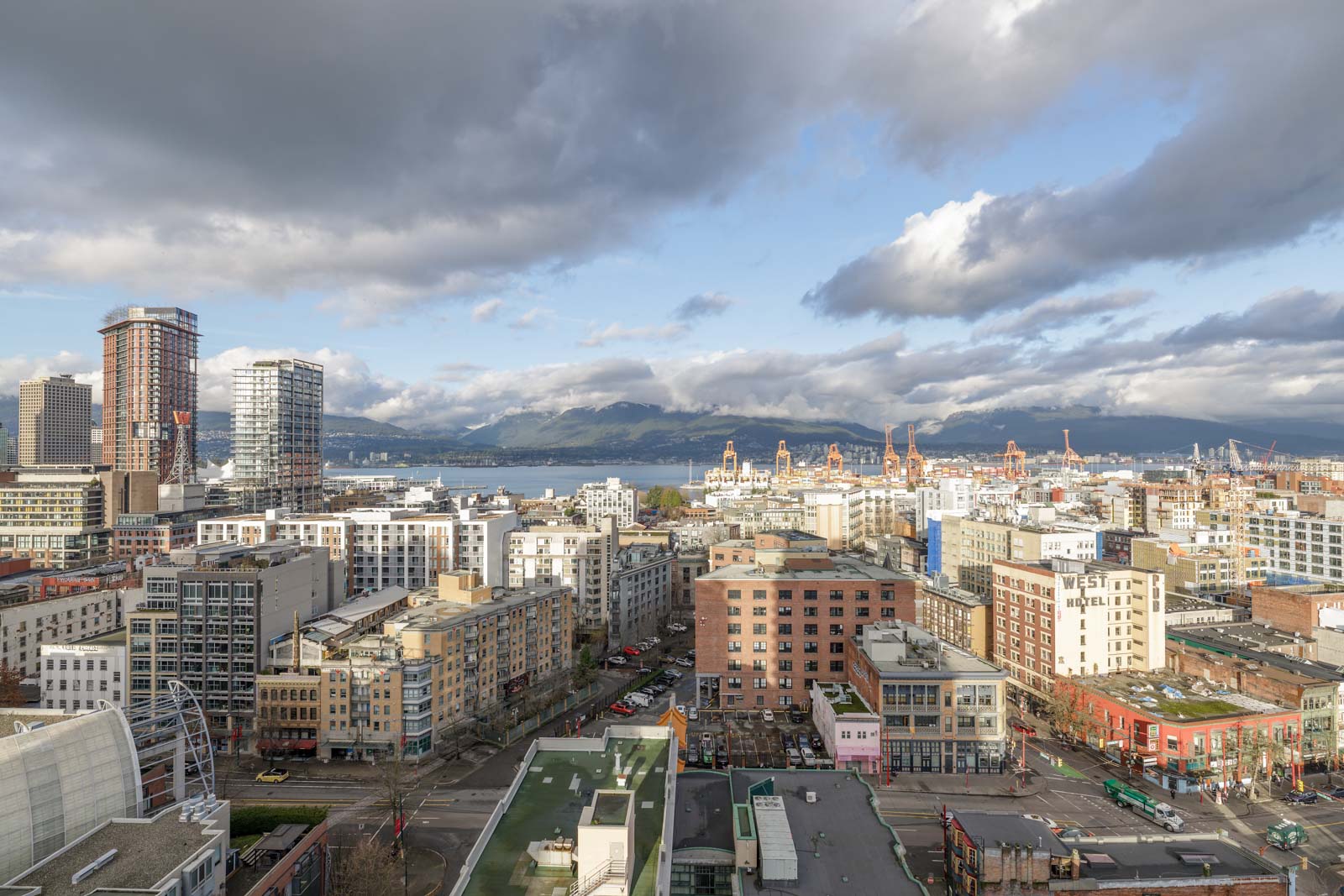 Cityscape view of a downtown area with numerous buildings and skyscrapers under a partly cloudy sky, with a distant view of mountains and water in the background.