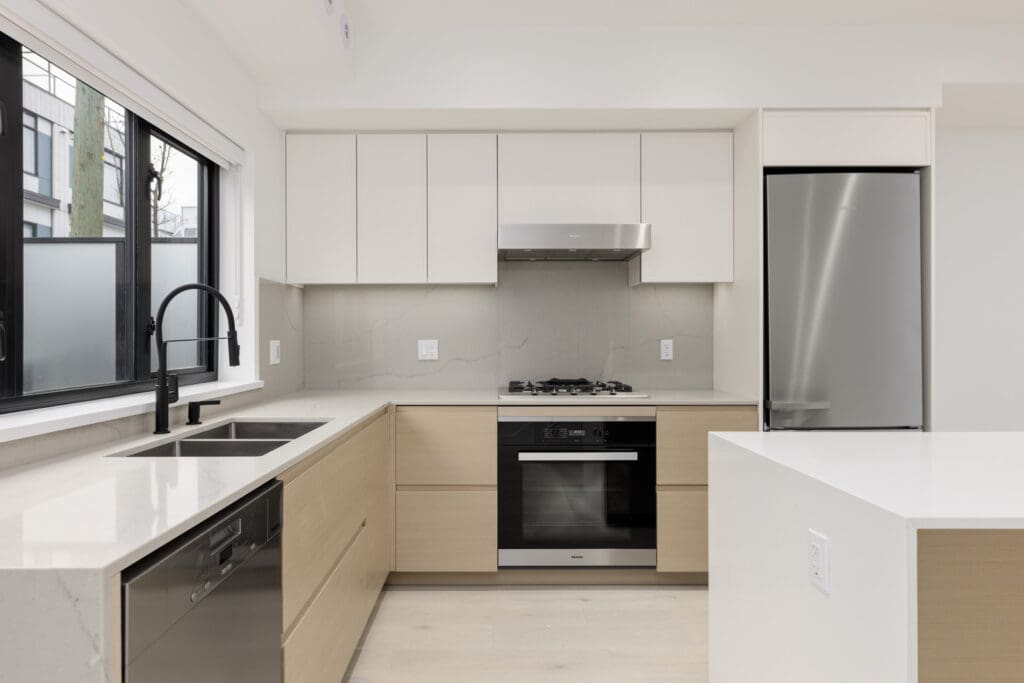 Modern kitchen with sleek white cabinets, stainless steel appliances, black faucet, and a large window. Features a built-in oven, gas stove, and light wood accents.
