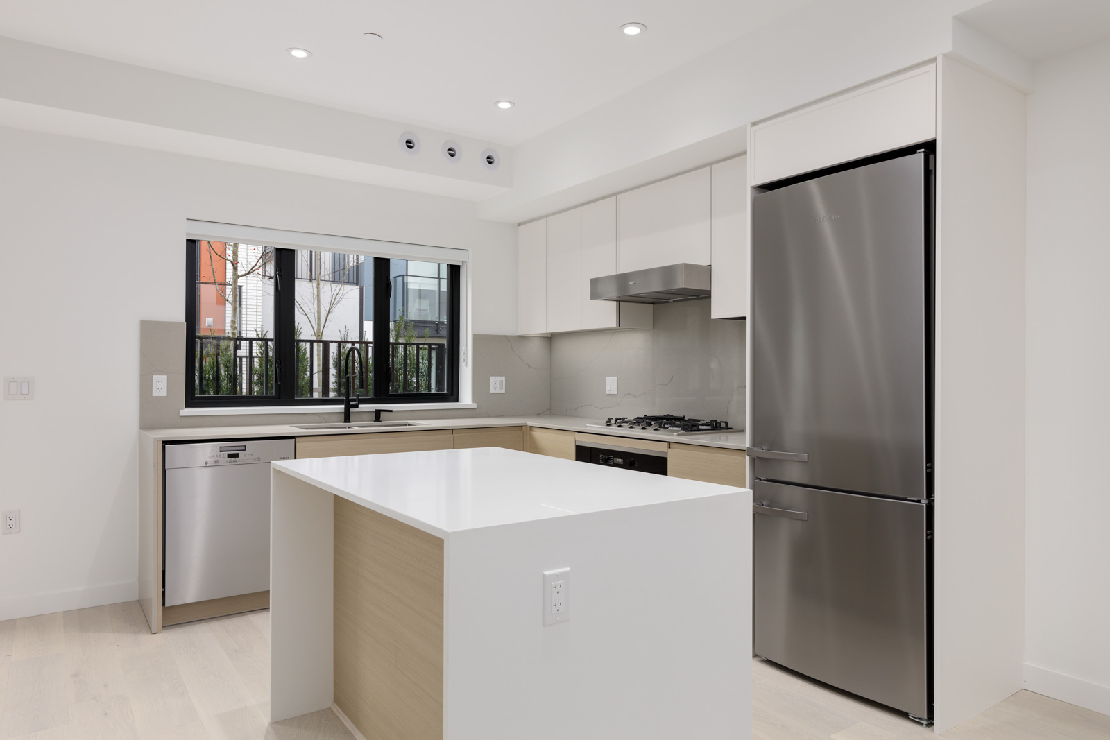 Modern kitchen with a central island, stainless steel appliances, and a window. White cabinets, light wood accents, and recessed lighting complement the sleek design.