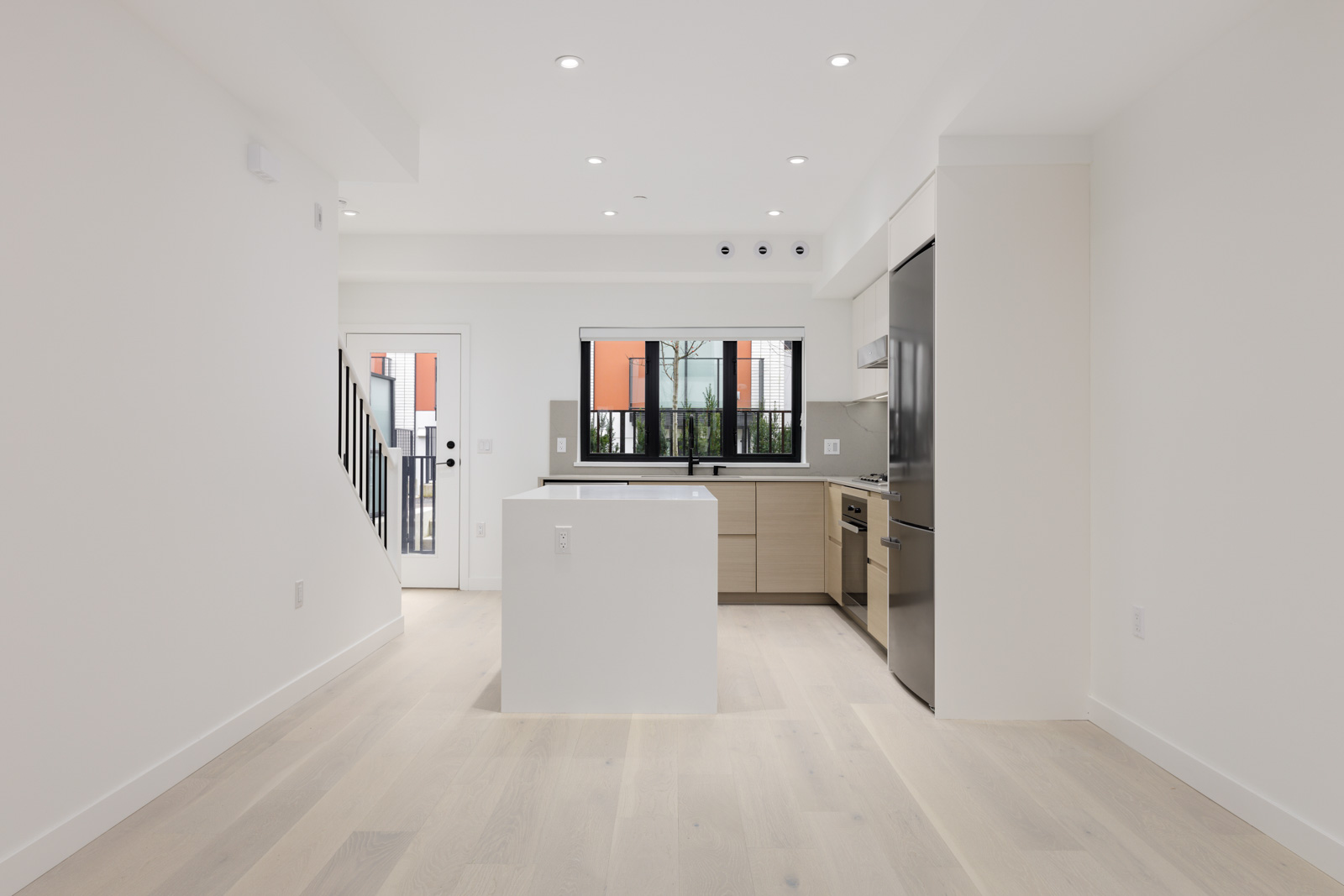Modern, bright kitchen with white walls, light wood flooring, stainless steel appliances, and a central island. Large window and glass door provide natural light.