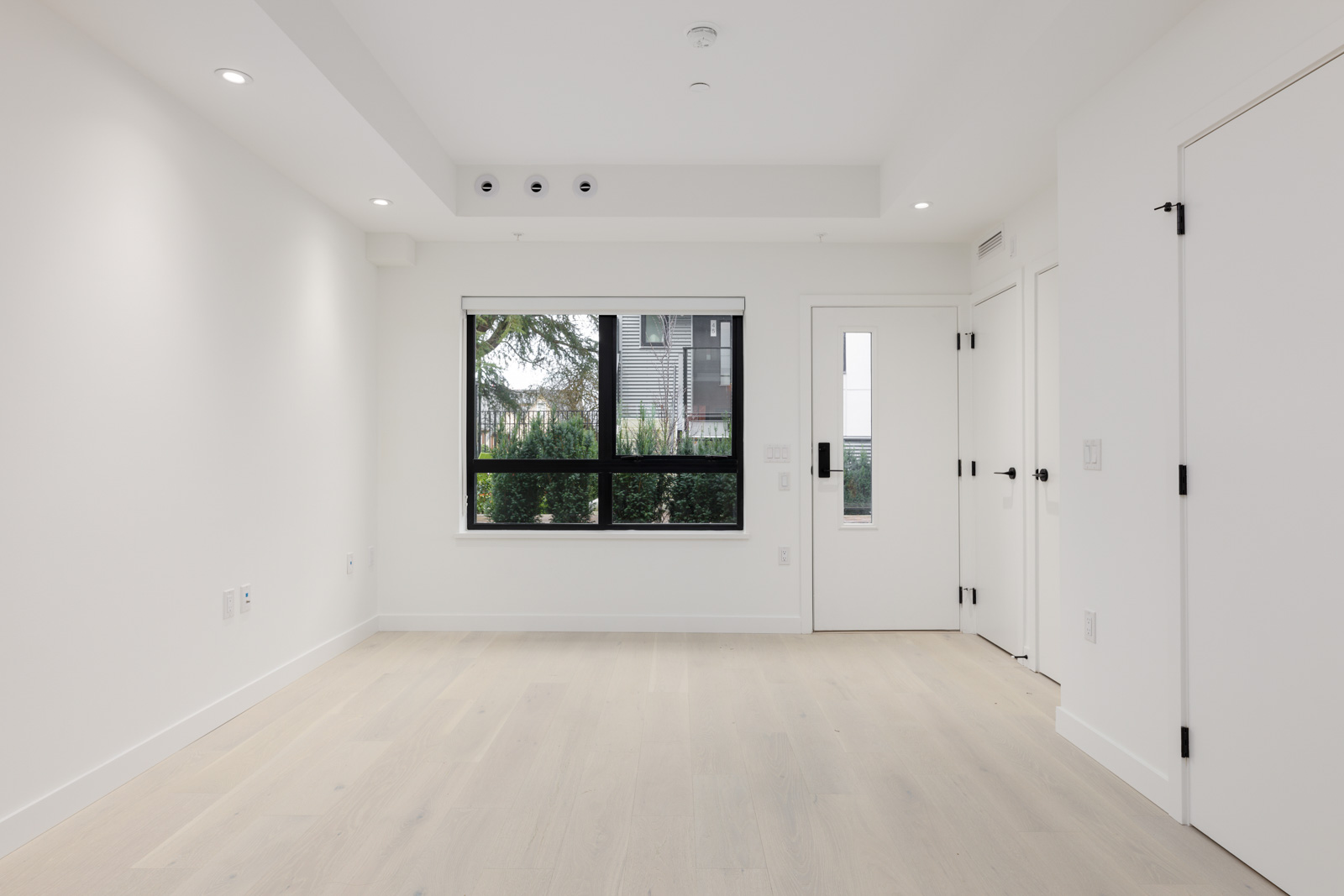 Empty white room with light wood flooring, recessed lighting, a large window, and a door. Outdoor view includes greenery and a building.