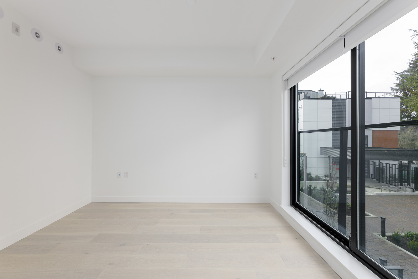 Empty white room with light wood flooring, large window showing an outdoor view with buildings and trees.