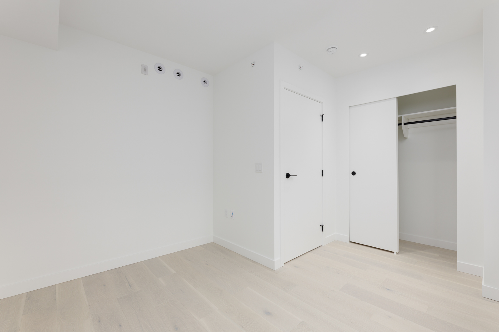 Empty room with light wood flooring, white walls, and a closed door. A sliding door closet with an open panel is on the right. Ceiling lights are on.
