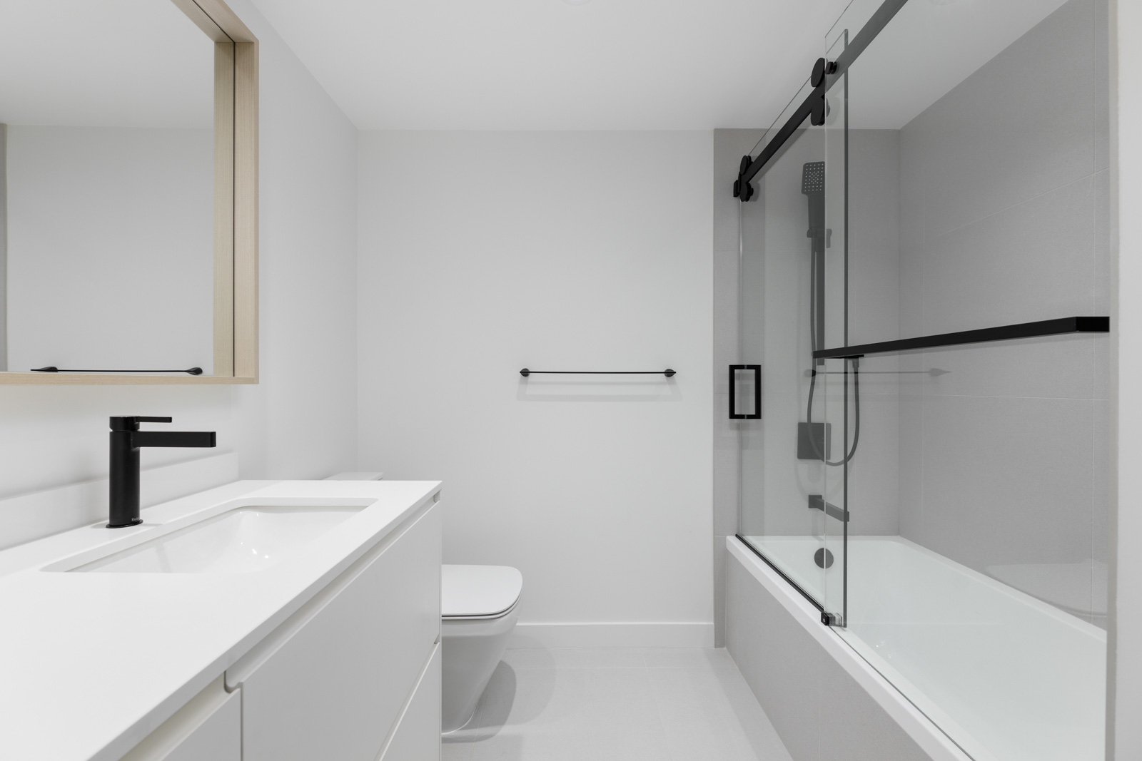 Modern bathroom with white walls, a vanity with a black faucet, a wall-mounted mirror, a toilet, and a bathtub with a glass sliding door.