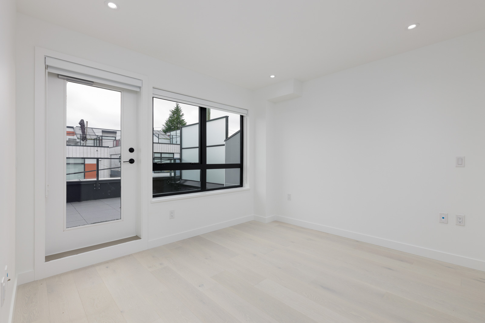Empty white room with light wood flooring, featuring a glass door leading to a balcony and a large window with a view outside.