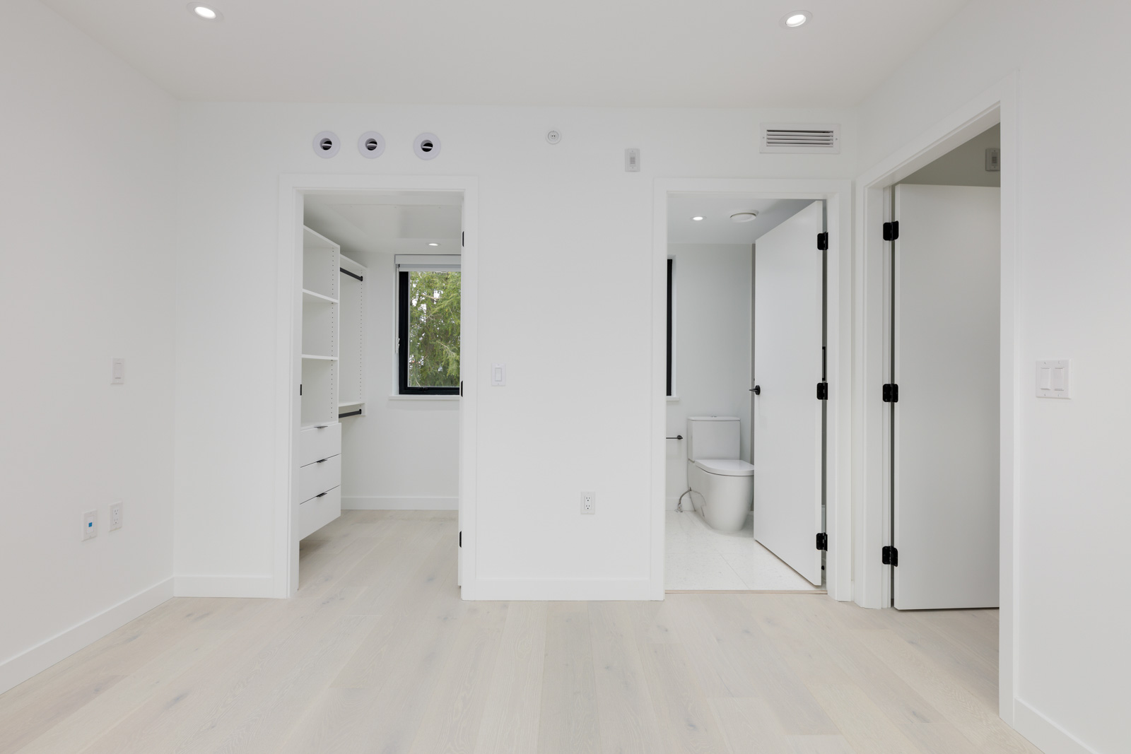 Minimalist white room with light wood floor, open closet with shelves, a door to a bathroom with a visible toilet, and a window with a view of trees.