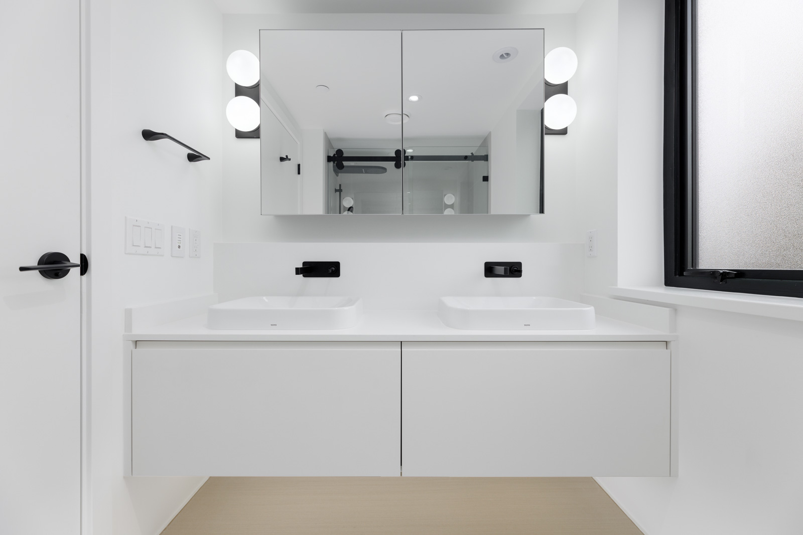 Modern bathroom with a double sink vanity, wall-mounted black fixtures, mirrored cabinet, and frosted window. Walls and surfaces are white, creating a minimalist design.