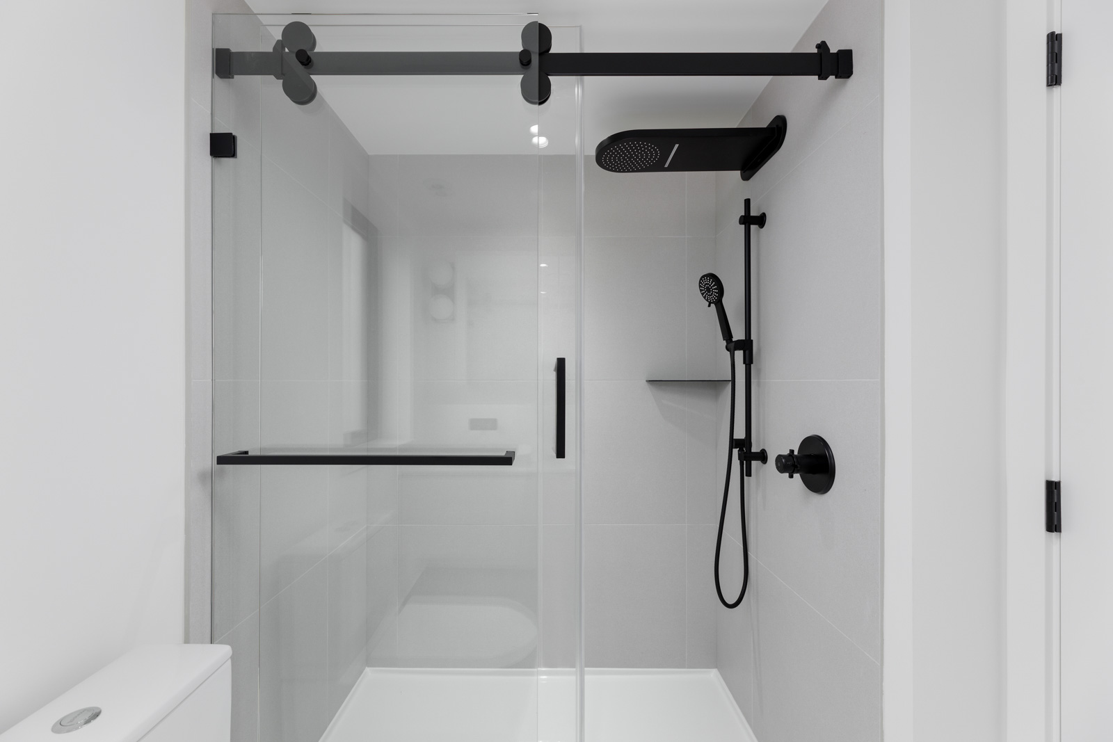 Minimalist shower with glass sliding door, black fixtures, and handheld showerhead in a white-tiled bathroom.