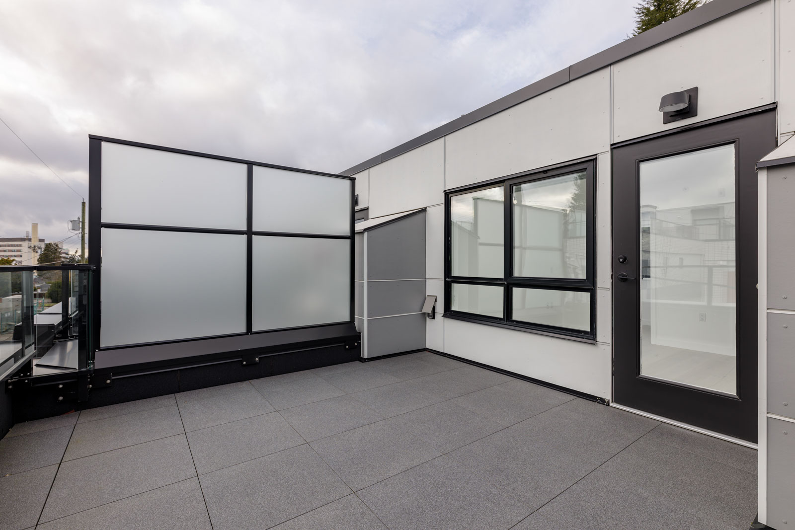 Modern rooftop patio with gray tiles, frosted glass privacy panels, and large windows. Overcast sky above.