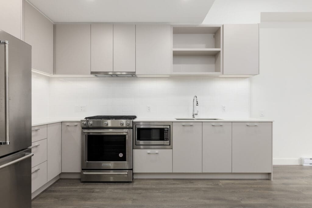 Modern kitchen with stainless steel appliances, including a refrigerator, oven, and microwave. White cabinets, light countertops, and wood flooring enhance the minimalist design.