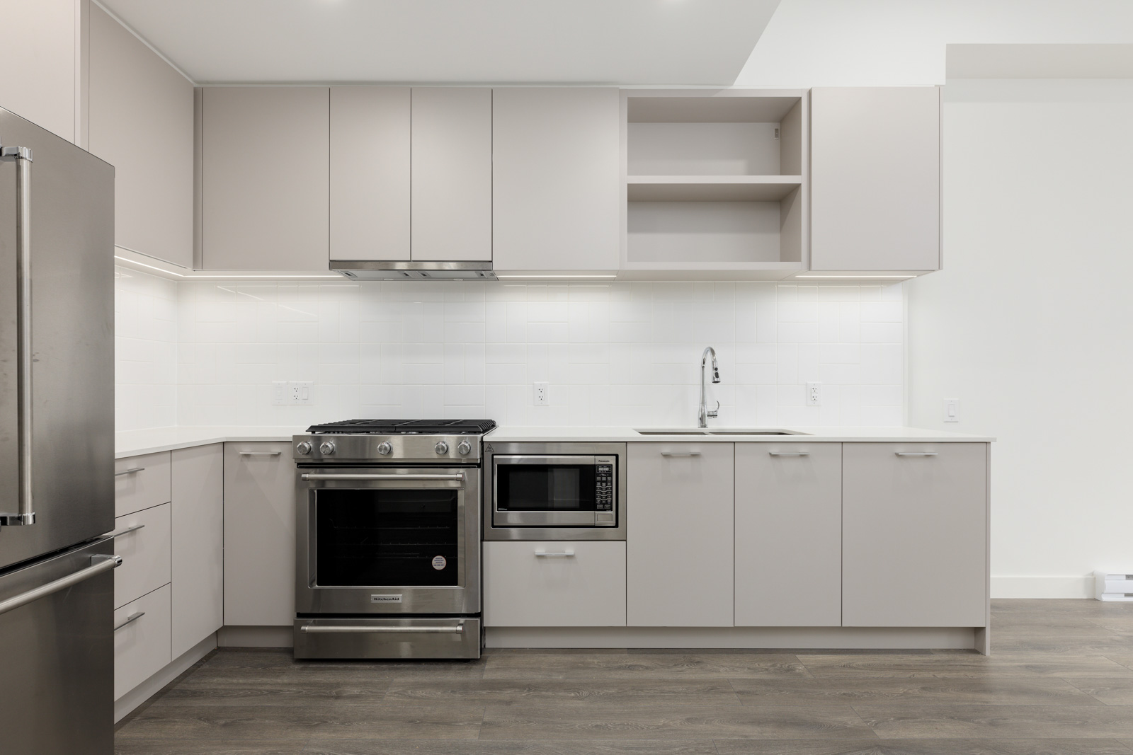 Modern kitchen with stainless steel appliances, including a refrigerator, oven, and microwave. White cabinets, light countertops, and wood flooring enhance the minimalist design.