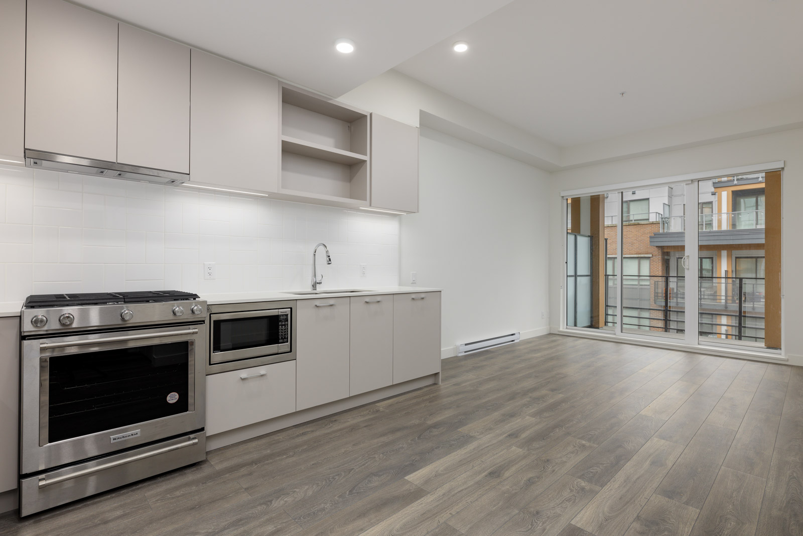 Modern kitchen with light-colored cabinets, stove, microwave, and sink. Hardwood floors and large sliding glass doors leading to a balcony with views of neighboring buildings.