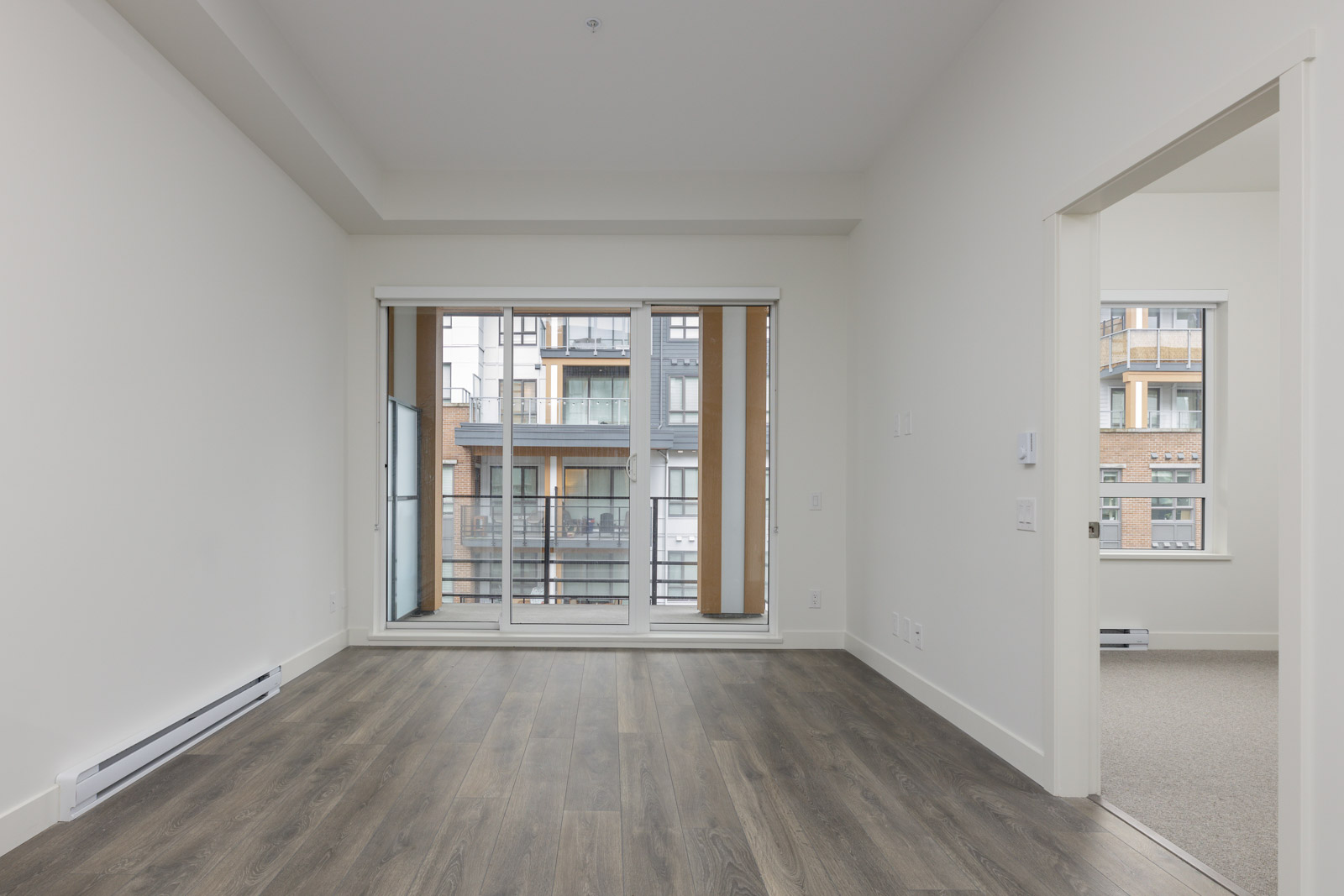 An empty room with hardwood floors, a large window with vertical blinds, and a view of an apartment building. An open doorway leads to a carpeted area.