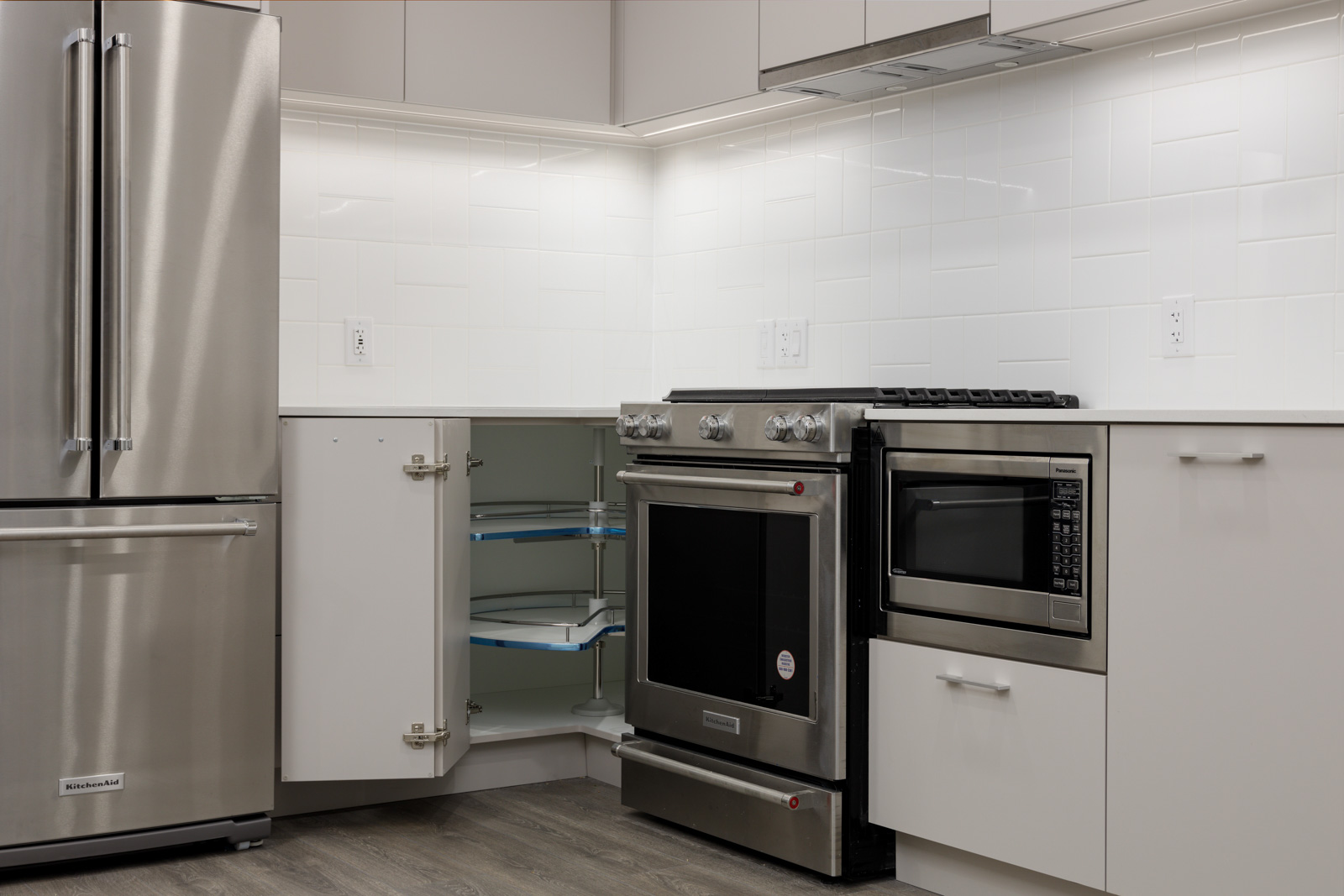 Modern kitchen with stainless steel fridge, oven, and microwave. White cabinets and tiled backsplash. Open corner cabinet with pull-out shelves. Wood flooring.