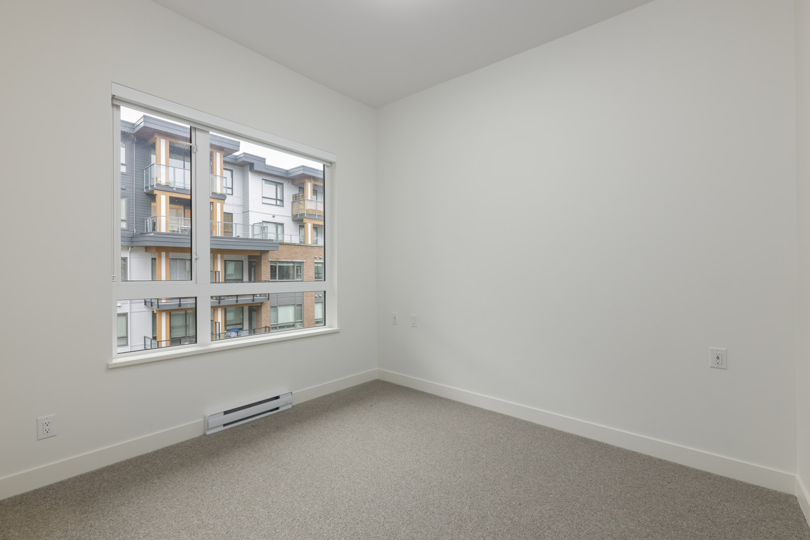 Empty room with beige carpet and white walls, featuring a large window overlooking an apartment building.