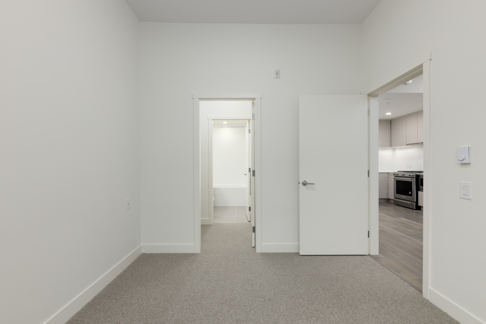 Empty room with carpeted floor, white walls, open doorways to a bathroom and a kitchen. The kitchen has wooden flooring and visible appliances.